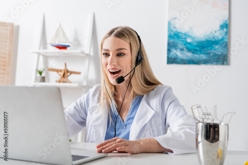Cheerful speech therapist in headset having video call on blurred laptop near tools in consulting room. © LIGHTFIELD STUDIOS