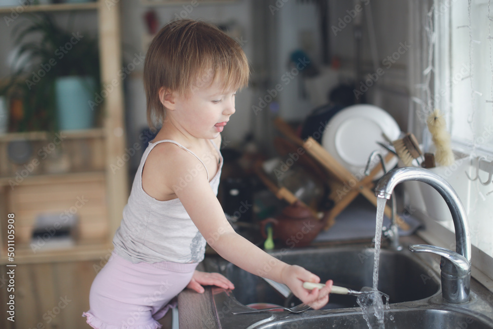 Toddler child washes his hands and fork in the sink in the kitchen, lifestyle in a real interior Toddler child washes his hands and fork in the sink in the kitchen, lifestyle in a real interior