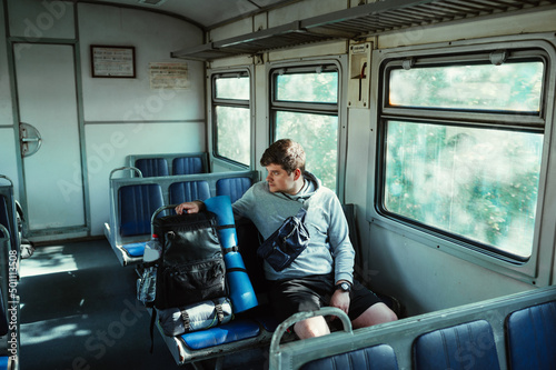 Male tourist in casual clothes rides in a train sitting on a bench with a backpack and goes hiking.