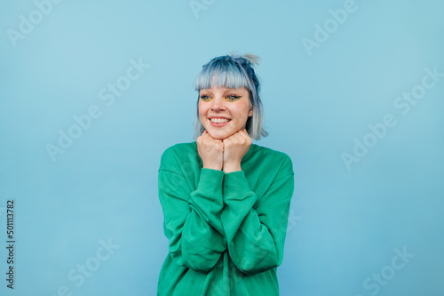 Happy hipster girl with colored hair stands on a blue background with a smile on his face and joyful looks away.