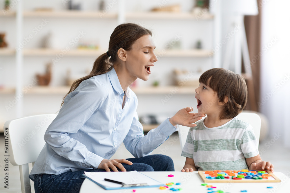 Fototapeta premium Professional woman speech therapist helping little boy to pronounce right sounds, showing mouth articulation
