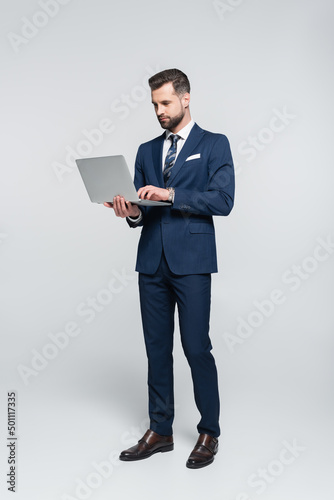 full length view of businessman in blue suit using laptop on grey.