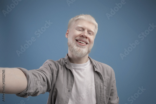 Positive unusual albino man taking selfie on turquoise background, cheerfully smiling at camera