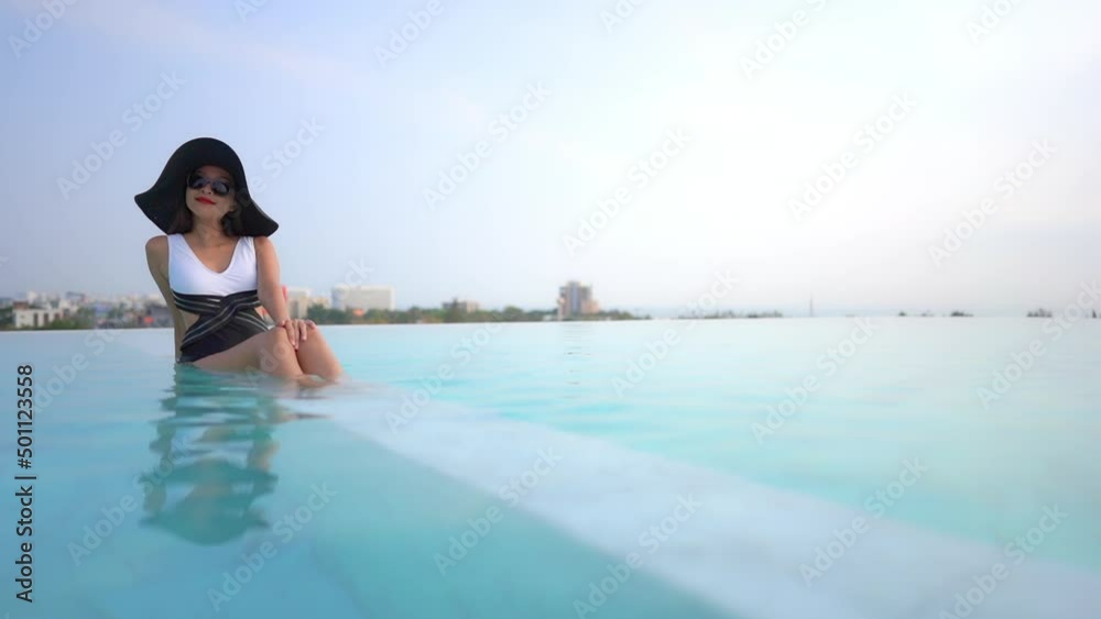 Lady in swimsuit and large hat relaxing in warm clear swimming pool with view of buildings, landscape and sea in background. Asian woman on vacation sitting in infinity pool tanning. Lady in swimsuit and large hat relaxing in warm clear swimming pool with view of buildings, landscape and sea in background. Asian woman on vacation sitting in infinity pool tanning.