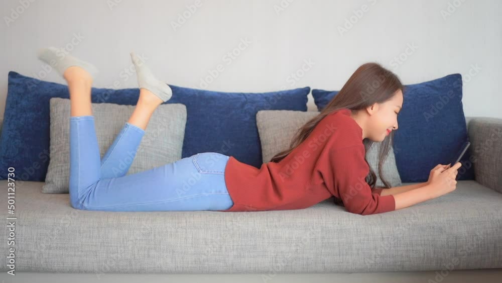 Young woman laying on stomach using smartphone in living room on sofa checking online profile or dating app. Attractive Asian lady texting, gaming, gambling or shopping on phone. Slow motion. Young woman laying on stomach using smartphone in living room on sofa checking online profile or dating app. Attractive Asian lady texting, gaming, gambling or shopping on phone. Slow motion.
