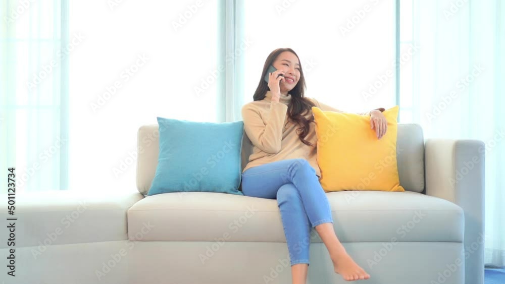 Slow motion view of young Asian woman seated on sofa near bright window answers call on cell phone, smiles and talks. Happy Chinese lady in living room talking on phone. Slow motion view of young Asian woman seated on sofa near bright window answers call on cell phone, smiles and talks. Happy Chinese lady in living room talking on phone.