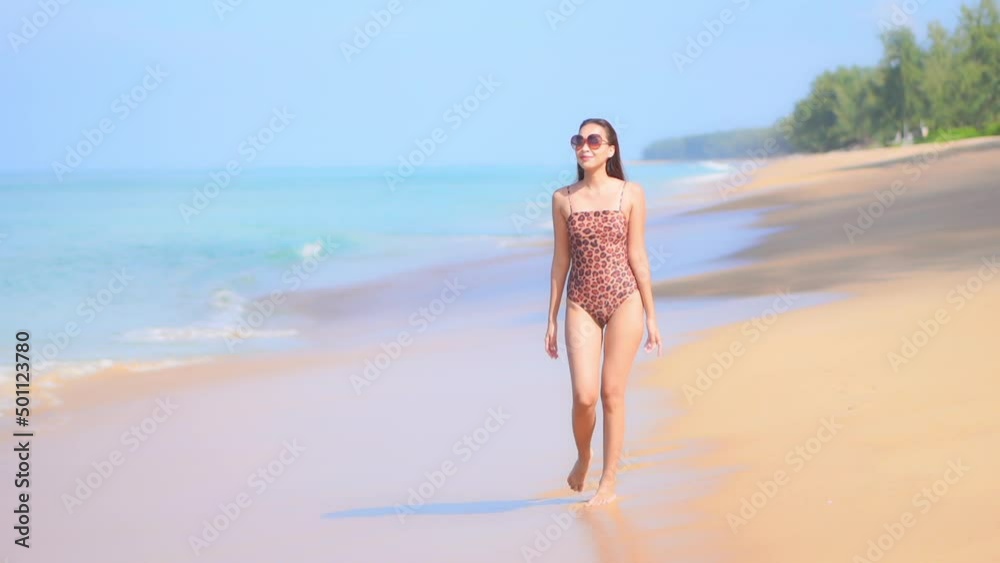 Slow motion view of attractive Asian woman in swimsuit walking alone on beach with waves of water rushing onto shore. Tropical island paradise vacation holiday destination. Lady strolling seaside. Slow motion view of attractive Asian woman in swimsuit walking alone on beach with waves of water rushing onto shore. Tropical island paradise vacation holiday destination. Lady strolling seaside.