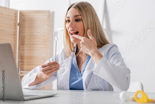 Speech therapist holding tool and pointing at mouth during video call on laptop.