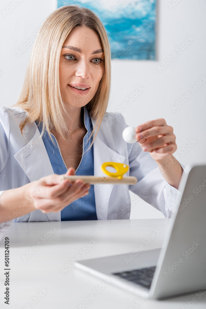Positive logopedist holding respiratory muscle trainer during video call on laptop in consulting room. Positive logopedist holding respiratory muscle trainer during video call on laptop in consulting room.