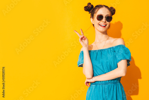 Young beautiful smiling female in trendy summer jeans overalls. Sexy carefree woman with two horns hairstyle posing near yellow wall in studio. Positive model having fun. Isolated. In sunglasses