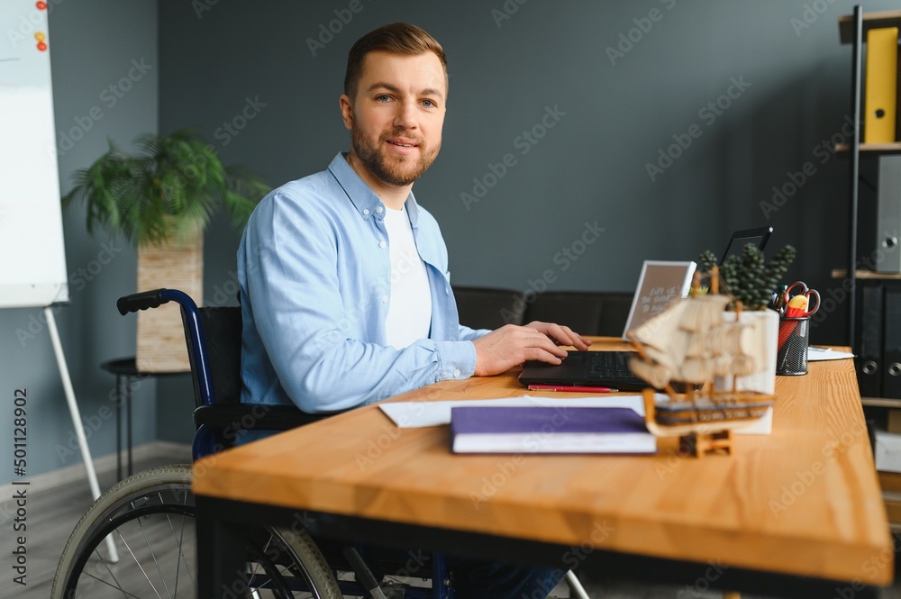 Young man with special needs in casual clothes working on wireless laptop. Male freelancer working from home while sitting in wheelchair.