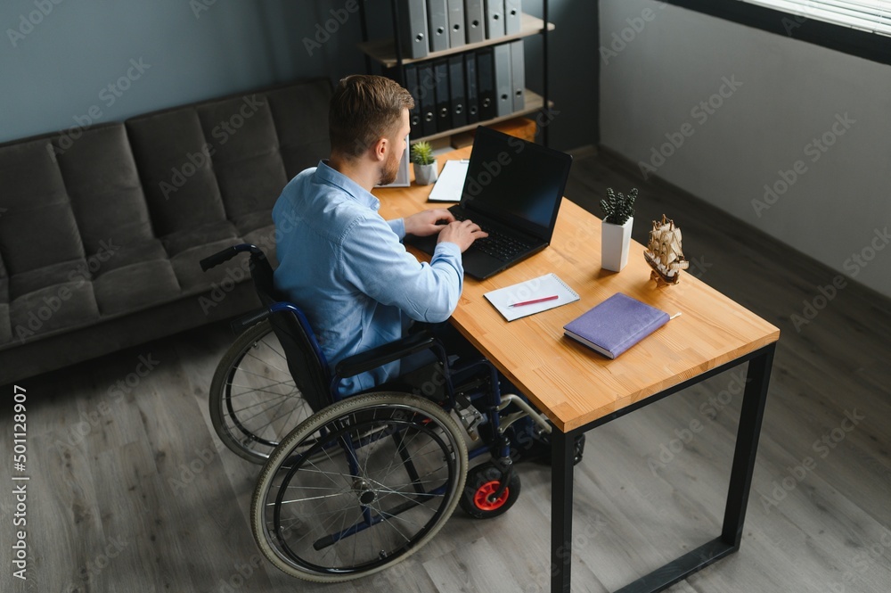 Young man with special needs in casual clothes working on wireless laptop. Male freelancer working from home while sitting in wheelchair. Young man with special needs in casual clothes working on wireless laptop. Male freelancer working from home while sitting in wheelchair.