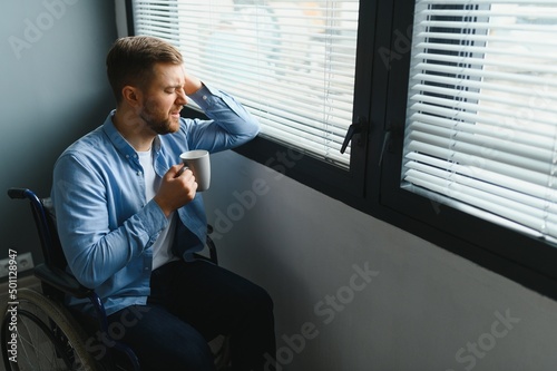 Disabled Person Sits in Wheelchair Against Window. Serious Sad Caucasian Man Wearing Casual Clothes and Look at Large Panoramic View in Bright Modern Living Room or Hospital.