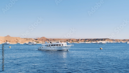 Large white yacht sailing across blue sea on sunny day, against marina backdrop with anchored yachts. Luxury motor yacht side view emerging from ship dock. Egypt Red Sea Cruise. Holiday yacht vacation
