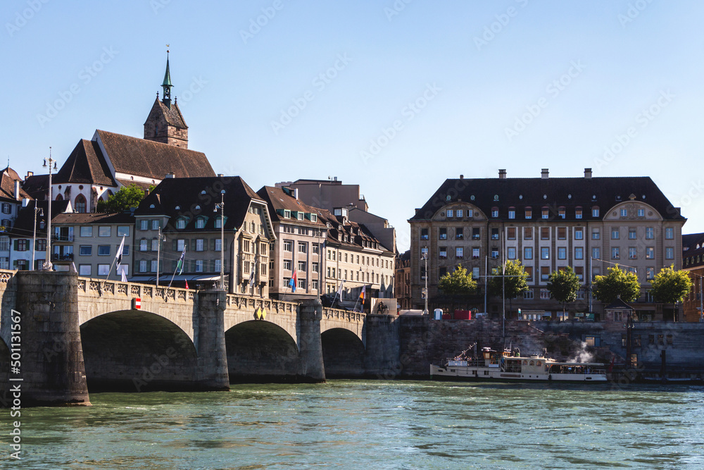 Fototapeta premium View of the old town of Basel, Switzerland, with an ancient stone bridge over the Rhine river and historical buildings and a church with a high sharp spire, a tourist ship at the pier