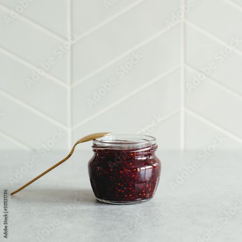 Small glass jar with ready to eat raspberry jam and golden spoon against grey kitchen tile wall with copy space. Natural window light, soft selective focus.