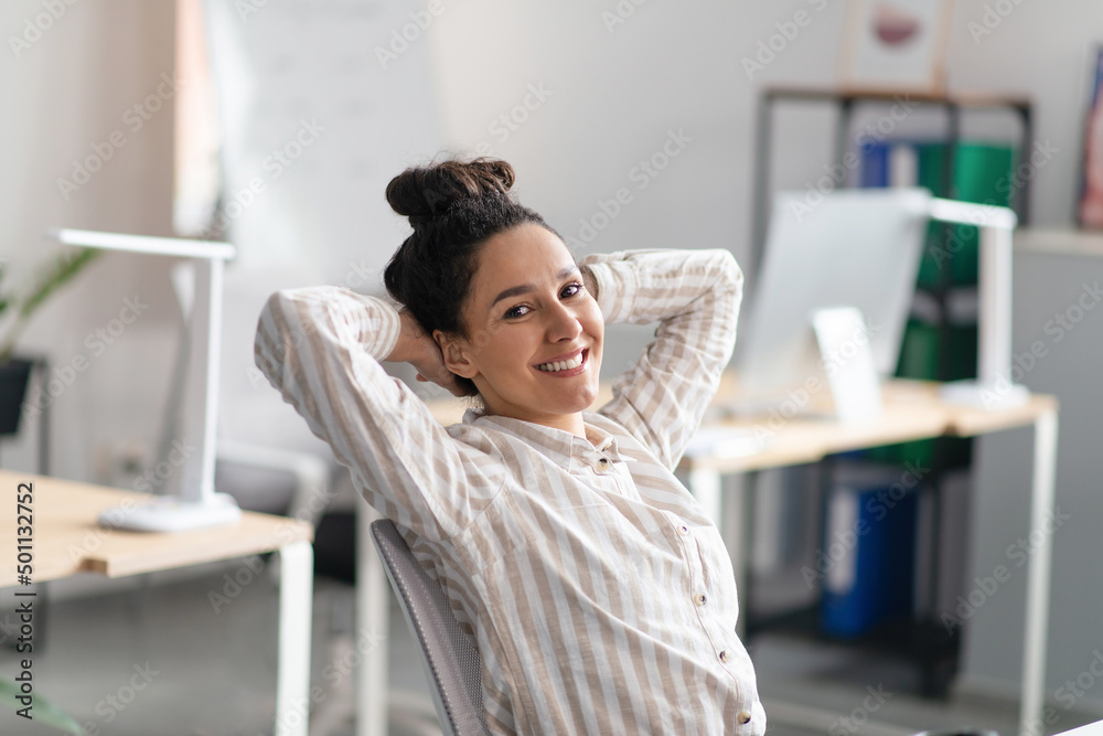 Happy relaxed female with hands behind her head enjoying rest from work in office interior, sitting at workplace Happy relaxed female with hands behind her head enjoying rest from work in office interior, sitting at workplace