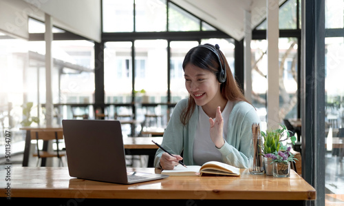 Happy young woman in headphones speaking looking at laptop making notes, girl student talking by video conference call, female teacher trainer tutoring by webcam, online training, e-coaching concept © wichayada