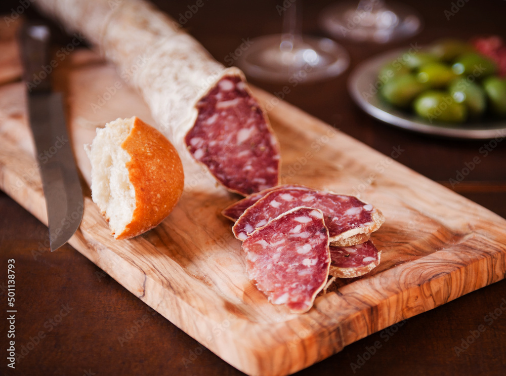 Salami Sliced on a Cutting Board with Bread; Bowl of Green Olives in the Background. Salami Sliced on a Cutting Board with Bread; Bowl of Green Olives in the Background.