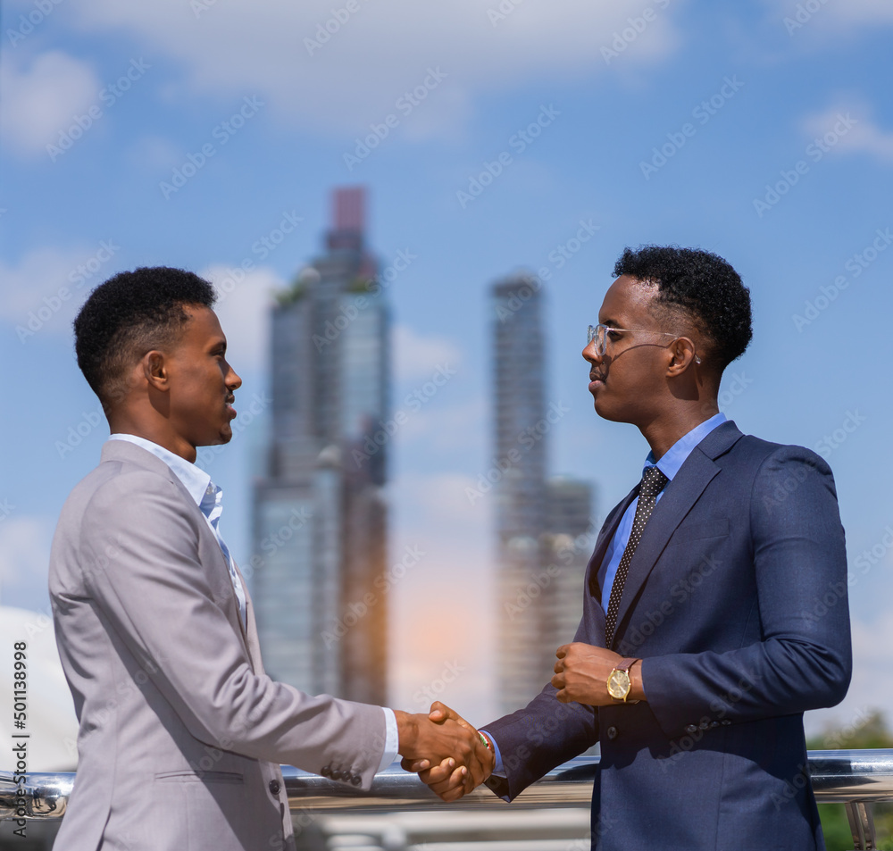 businessmen hand shake together, building in city with light at background. black man in grey suit and executive man with glasses in blue suit businessmen hand shake together, building in city with light at background. black man in grey suit and executive man with glasses in blue suit
