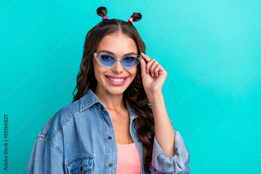 Photo of young adorable girl wear eyeglasses toothy smile bun hairdo isolated over teal color background Photo of young adorable girl wear eyeglasses toothy smile bun hairdo isolated over teal color background