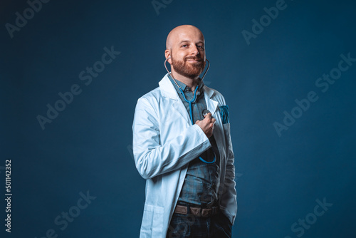 Photo of cheerful doctor with beard and stethoscope posing and smiling at camera with blue background and medical white coat