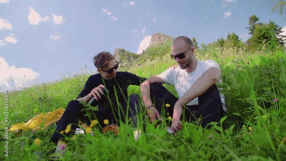 Two young men in dark glasses are sitting on a slope, chatting about life in the highlands. One guy supports the other. Tourism and travel Two young men in dark glasses are sitting on a slope, chatting about life in the highlands. One guy supports the other. Tourism and travel