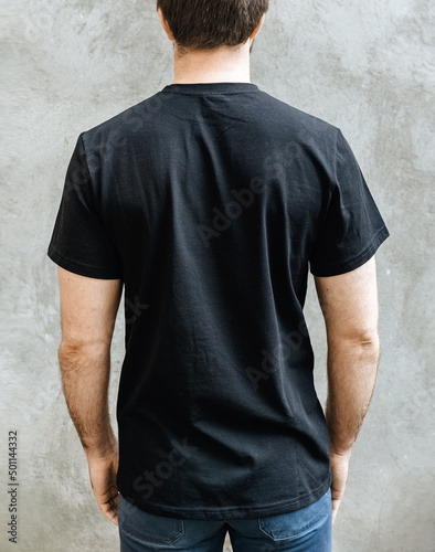 Young man in an empty black casual t-shirt. Rear view on a background of light gray concrete wall. Design and layout of men's t-shirt for printing.