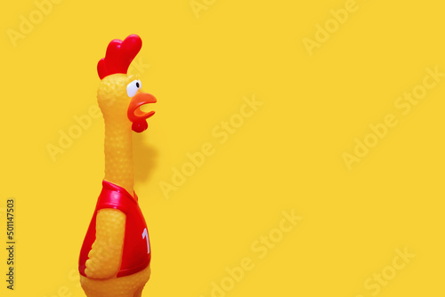 Rubber toy in the form of a rooster on a yellow background. The funny toy rooster has a surprised and dumbfounded look with its beak open. The toy makes loud noises. Free space for text and ads