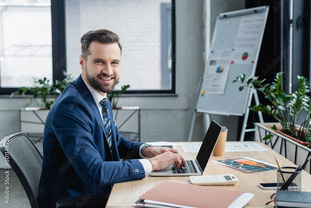 happy businessman looking at camera while typing on laptop with blank screen.