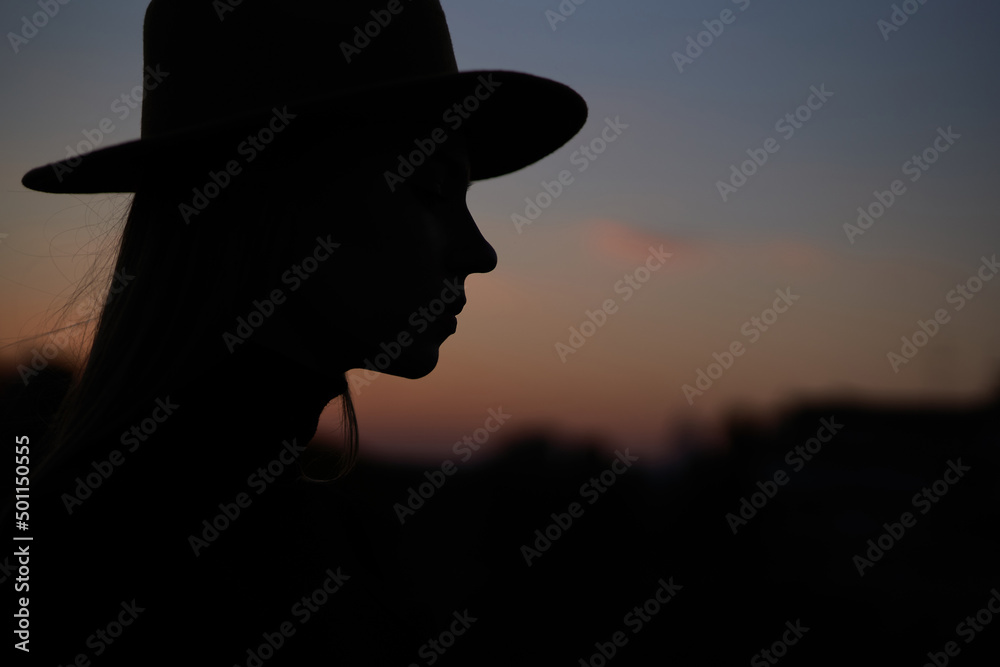 Closeup silhouette portrait of happy fashionable long hair woman wearing hat at sunset. Image against sun and blue sky. High quality horizontal photo