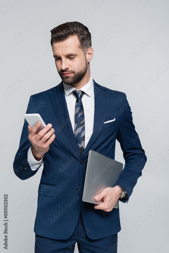 brunette businessman with laptop looking at mobile phone isolated on grey. brunette businessman with laptop looking at mobile phone isolated on grey.
