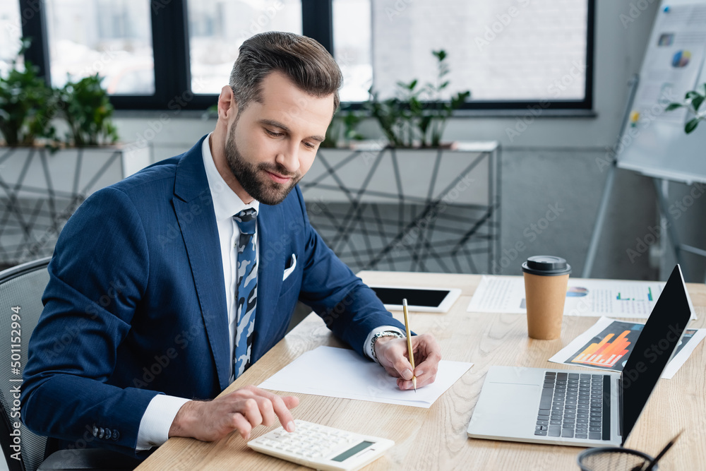 accountant with pencil using calculator while working near laptop in office. accountant with pencil using calculator while working near laptop in office.