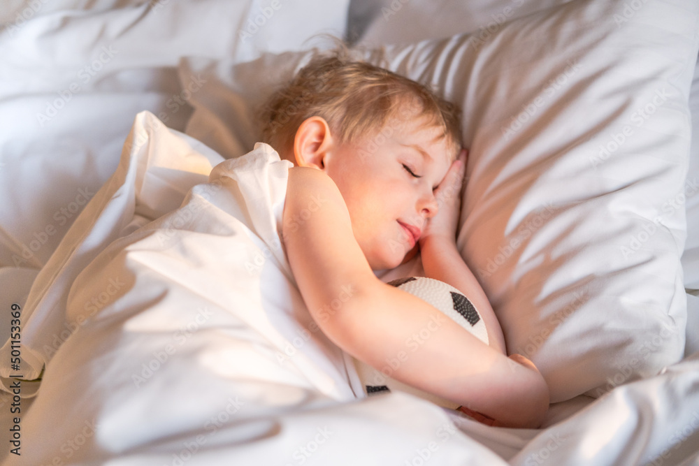 Obraz premium Toddler boy sleeping with a soccer ball on a white bed linen.