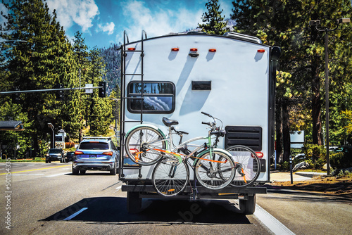 Camper with two bicycles tied to back on tree-lined highway outside of Lake Tahoe with mountains in distance on sunny summer da