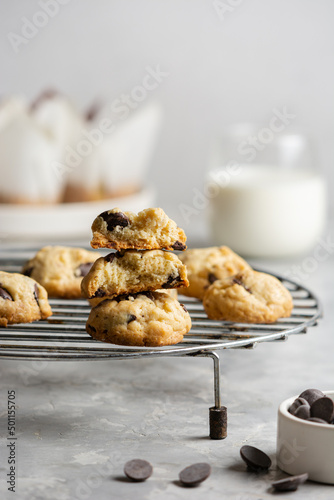 Chocolate chip cookies on a cooking lattice on a light gray concrete background, with a glass of milk, a plate of muffins and a bowl of chocolate drops