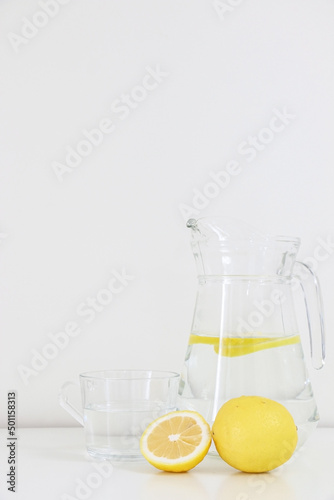 Bright image of refreshing healthy natural citrus lemon water, lemonade, in glass jug and cup besides a full and sliced lemon fruit, on white table against white wall background