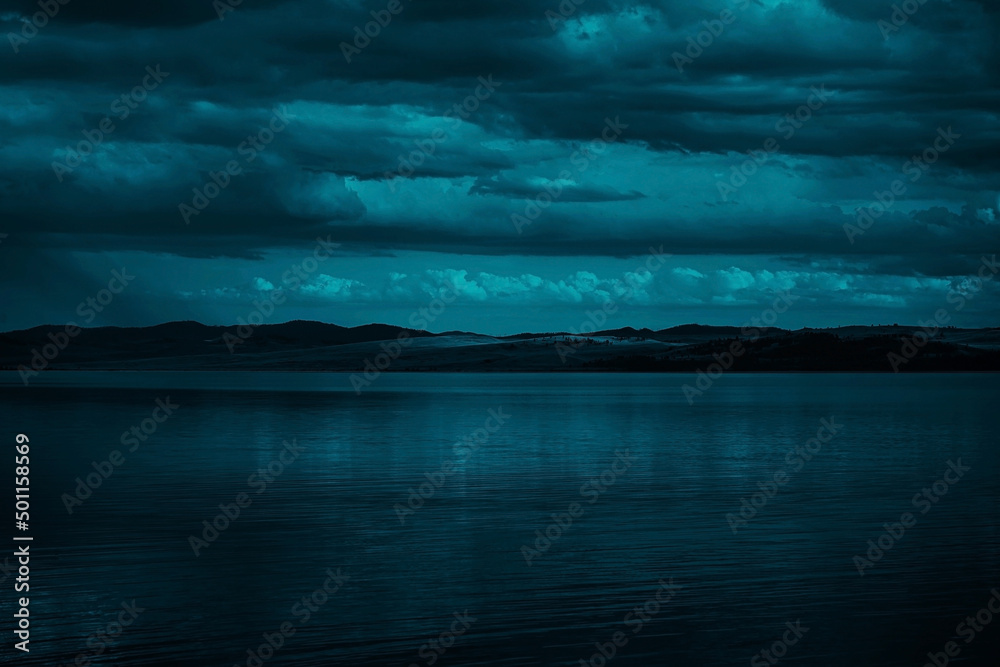 Blue-green clouds over the sea. Toned dark water and sky. Background with space for design. Silhouette of mountains on the horizon. Calm atmosphere. Moonlight. Fantasy, mystic.