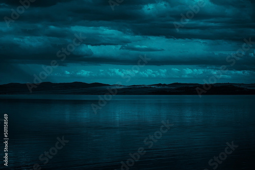 Obraz na płótnie Blue-green clouds over the sea. Toned dark water and sky. Background with space for design. Silhouette of mountains on the horizon. Calm atmosphere. Moonlight. Fantasy, mystic. w salonie