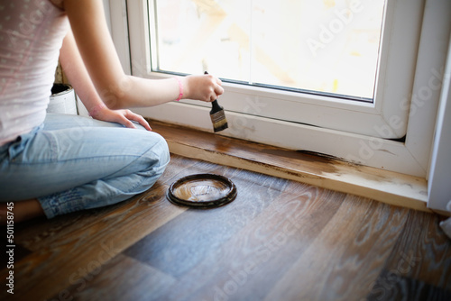 Kid girl with pigtails paints, covers a wooden board threshold with dark impregnation (oil), top view. Home renovation and creativity