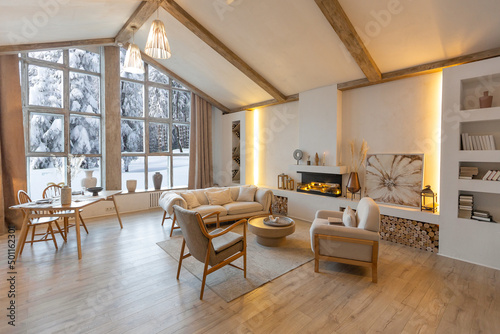 cozy warm home interior of a chic country chalet with a huge panoramic window overlooking the winter forest. open plan, wood decoration, warm colors and a family hearth