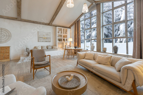 cozy warm home interior of a chic country chalet with a huge panoramic window overlooking the winter forest. open plan, wood decoration, warm colors and a family hearth