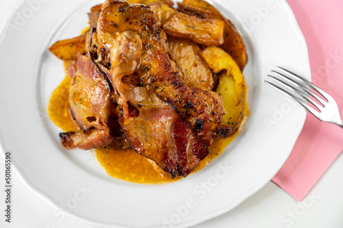 Grilled pork neck steak, bacon, baked potato on white plate, fork, pink towel on white.