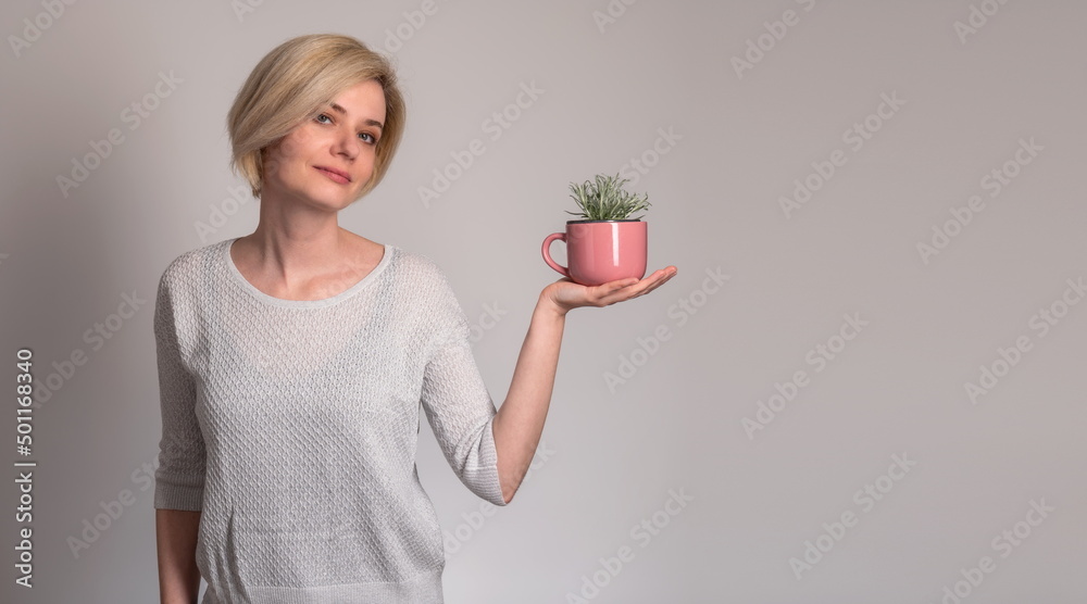 Portrait of stylish, nice, cute, modern woman in denim, casual outfit having empty place, copy space, looking at camera, holding a pink cup with a plant growing, on grey background. Concept: herb tea
