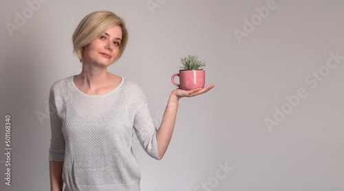 Obraz na płótnie Portrait of stylish, nice, cute, modern woman in denim, casual outfit having empty place, copy space, looking at camera, holding a pink cup with a plant growing, on grey background. Concept: herb tea w salonie