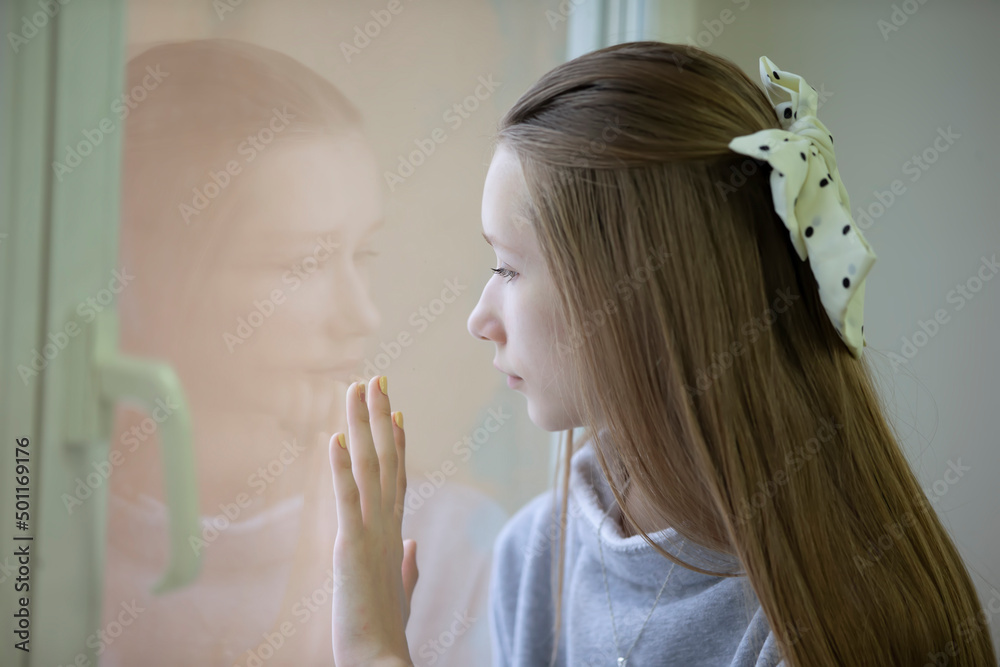Fototapeta premium Sad teenager girl looks out the window, feels lonely. Thoughtful girl is unhappy with the problem. Difficult adolescence concept.