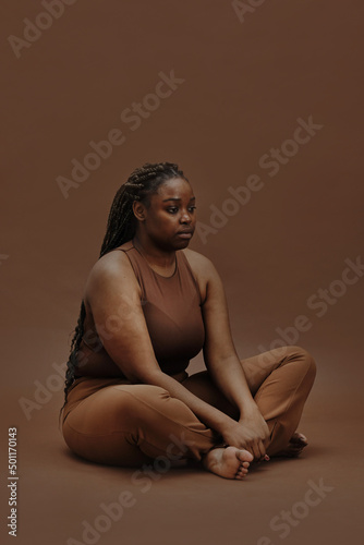 Portrait of young black woman in casual clothing sitting with blank expression isolated on brown background