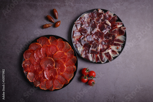 Portions of acorn-fed Iberian loin and 100% Dehesa de Extremadura acorn-fed Iberian ham on a black plate and gray table, decorated with tomatoes and acorns photo