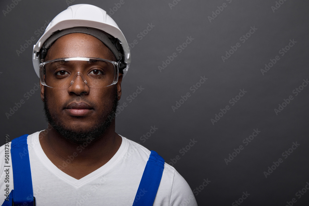 Close-up portrait of a handsome serious locksmith in a white helmet on a gray wall. In a white T-shirt and overalls with blue straps poses a young worker in construction goggles Close-up portrait of a handsome serious locksmith in a white helmet on a gray wall. In a white T-shirt and overalls with blue straps poses a young worker in construction goggles