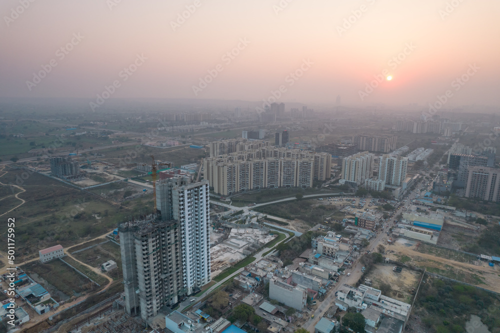aerial drone shot passing over a building with homes, offices, shopping centers moving towards skyscapers in front of sunset showing the empty outskirts of the city of gurgaon